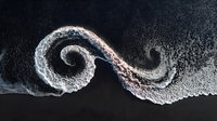 Dramatic aerial view of ocean waves forming a spiral pattern against dark shoreline, creating natural abstract art for coastal home decor.