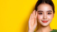 Young Asian woman with natural makeup listening carefully with hand near ear against vibrant yellow background, expressing curiosity and attention.
