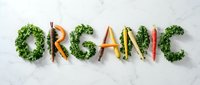 Word organic spelled with fresh colorful carrots and kale leaves on white marble surface, promoting healthy eating and natural food concepts.
