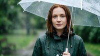 Young woman with reddish-brown hair holding transparent umbrella in rain, wearing dark jacket outdoors in green garden setting, suitable for weather, lifestyle and seasonal content.