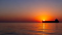Cargo ship silhouette sailing across calm ocean waters during golden sunset with vibrant orange and purple sky reflecting on sea surface for travel concepts.