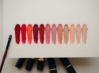 Lipstick swatches in a gradient from bold red to nude pink displayed on white surface with makeup brush and cosmetic tubes below.