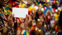 Hands holding blank white card at festive carnival celebration with colorful bokeh lights and costumed crowd in background, suitable for event marketing and promotional messaging.