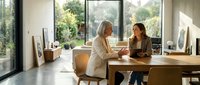 Two women having a professional meeting at a wooden desk in a bright, modern office space with large windows overlooking greenery, suitable for business, corporate, and workplace concepts.