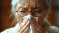 Senior Asian woman in glasses blowing nose into tissue during cold or flu illness, showing genuine emotional discomfort against warm background.