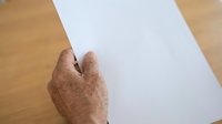Mature hands with age spots holding blank white paper on wooden surface for writing, printing, or document presentation purposes.