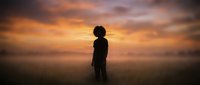 Young African American boy standing alone in misty field during golden hour sunset with dramatic orange sky clouds creating peaceful solitude moment.