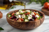 Fresh Mediterranean salad with feta cheese, olives and tomatoes in wooden bowl being drizzled with olive oil on marble counter for healthy eating.