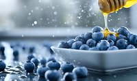 Fresh ripe blueberries in white ceramic bowl with golden honey dripping from wooden dipper, soft bokeh background creates dreamy atmosphere.