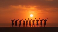 Silhouettes of diverse people with raised arms celebrating against dramatic orange sunset sky over ocean horizon.