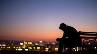 Young man sitting alone on bench during sunset overlooking city lights, contemplating life decisions and future opportunities in urban evening atmosphere.