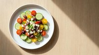 Fresh Greek salad with cherry tomatoes, cucumber, olives, and feta cheese on a white plate, top view, natural light on wooden table.