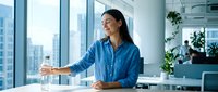 Young Asian businesswoman in blue shirt smiling while presenting in modern office with city skyline view. Professional corporate meeting environment.