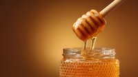 Golden honey dripping from wooden dipper onto honeycomb in glass jar against warm amber background, showcasing natural sweetness and viscosity.