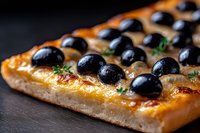 Homemade focaccia bread topped with black olives and fresh herbs on dark wooden surface, close up view showing texture and golden crust.
