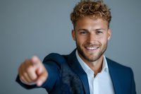 Young Caucasian businessman with curly blonde hair and beard wearing navy suit points forward with confident smile against gray studio background.