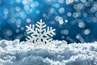 Decorative crystal snowflake ornament nestled in fresh snow against blurred blue bokeh background creates magical winter holiday atmosphere.