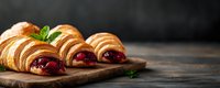 Fresh baked croissants filled with sweet raspberry jam on rustic wooden board, garnished with mint leaves against dark background, morning pastry.