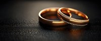 Golden wedding rings with diamonds on dark textured background with water drops, romantic symbol of marriage and eternal love in warm light.