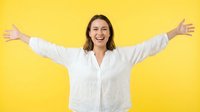 Woman with shoulder-length brown hair, Caucasian ethnicity, wearing white linen shirt, arms spread wide in celebratory gesture against bright yellow background. Suitable for success, happiness, freedom, and positive emotion concepts.