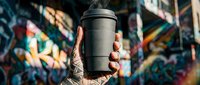 Tattooed hand holding black reusable coffee cup against vibrant street art graffiti wall background, eco-friendly lifestyle and sustainable beverage container concept.