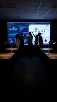 Business team analyzing data dashboard on large screen in dark conference room with silhouetted professionals discussing charts and graphs.