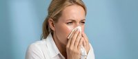 Woman with blonde hair, fair complexion, middle-aged, wearing white shirt, holding tissue to nose, expressing cold or allergy symptoms against blue background.