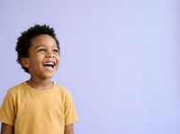 Young African American boy wearing mustard yellow t-shirt, smiling joyfully with head tilted upward against soft lavender background. Studio portrait suitable for parenting, education, and lifestyle marketing.