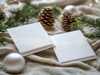 Blank white greeting cards mockup with pine cones, silver ornaments and fir branches on beige linen fabric. Holiday stationery photography setup.