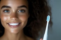 Young mixed race woman with bright smile and freckles holding electric toothbrush, demonstrating oral hygiene and dental care routine against dark background.