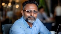 Middle-aged South Asian businessman wearing glasses and blue shirt smiling confidently in modern office setting. Professional portrait with blurred background.