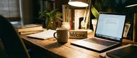Cozy home office desk with laptop, welcome sign, and coffee mug under warm lamp light, creating an inviting remote work atmosphere.