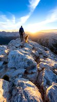 Male hiker and golden retriever dog standing on rocky mountain peak at sunset, dramatic sky and mountain range silhouettes create inspiring outdoor scene.