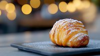 Fresh baked croissant dusted with powdered sugar on dark plate against blurred warm bokeh lights background, close up view of flaky pastry.