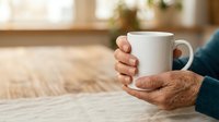 Senior man with brown skin holding white ceramic mug at wooden table indoors, wearing dark sweater. Warm beverage moment for lifestyle and wellness content.