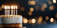Festive birthday cake with glowing candles against dark background with golden bokeh lights, partial view showing white frosting and caramel layer.