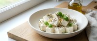 Fresh white cheese cubes with herbs and black pepper seasoning in ceramic bowl on wooden board, healthy Mediterranean appetizer ready to serve.