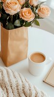 Peach roses in kraft paper bag alongside coffee cup and knitted throw blanket on white surface. Cozy morning lifestyle composition with soft natural lighting.