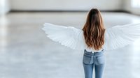 Young Caucasian woman with long brown hair wearing jeans and white angel wings standing in minimalist bright interior space, back view. Spiritual concept.