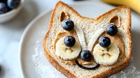 Playful cat-shaped toast with banana slices and blueberries creating cute animal face breakfast. Creative food art for children featuring fresh fruit on white bread.