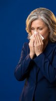 Mature woman with gray hair and Caucasian ethnicity wearing dark navy blazer blowing nose with tissue against solid blue background for healthcare and illness concepts.