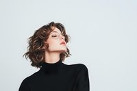 Young Caucasian woman with short wavy brown hair wearing black turtleneck and glasses poses confidently against light background, head tilted up with eyes closed in serene expression.