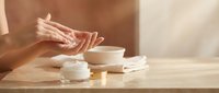 Woman applying moisturizer cream from jar on wooden table in soft natural light. Skincare routine, beauty treatment, cosmetic product application.