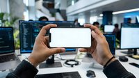 Person with dark skin tone holding smartphone with blank white screen in modern tech office workspace for app mockup and digital display purposes.