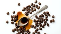 Roasted coffee beans scattered around golden cup with metal spoon on white background, suitable for cafe, beverage, and food industry marketing materials.