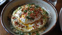 Creamy whipped labneh topped with runny poached egg, fresh herbs, olive oil, and spicy red pepper flakes served in rustic ceramic bowl on wooden surface.