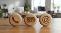 Three wooden circular tokens with engraved contact symbols including phone, email and at sign placed on office desk for business communication concept.