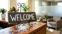 Rustic wooden welcome sign with decorative metal stand on reception desk in modern office waiting room with plants and comfortable seating area.