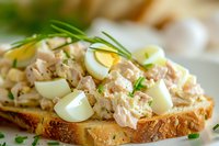 Creamy tuna salad sandwich with boiled egg quarters and fresh chives on toasted bread, close-up view showing texture and layers of homestyle preparation.