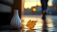White ceramic vase and fallen autumn leaf on wooden surface at sunset, with blurred walking person and golden bokeh lights creating atmospheric mood.